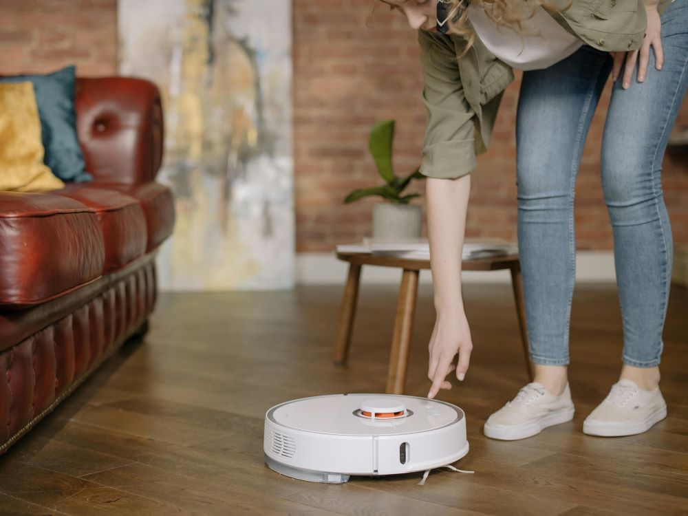Robot vacuum A person is about to press a button on a white robot vacuum cleaner on a wooden floor, with a red couch and a wooden table in the background.