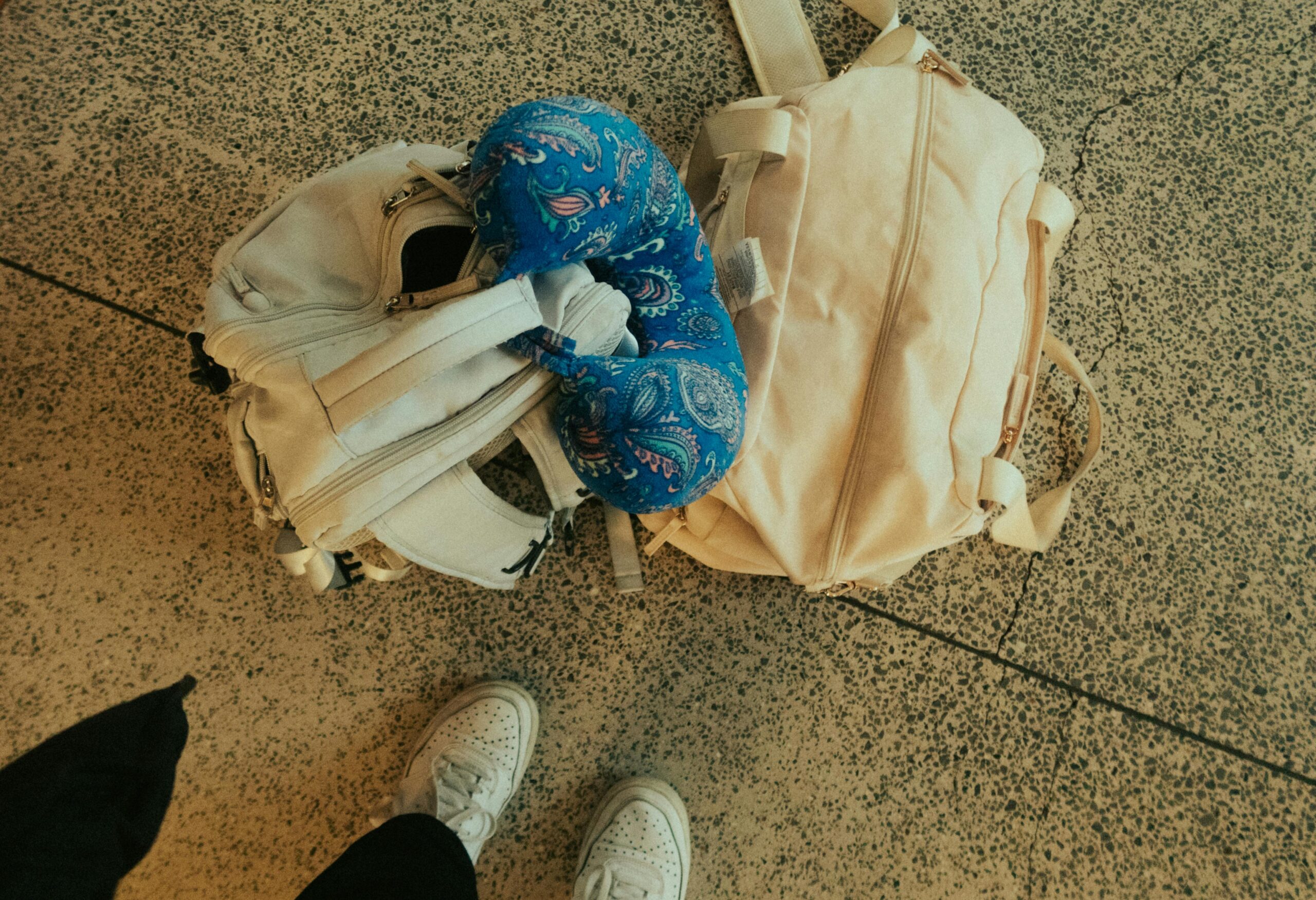 travelling light Two white bags and a blue patterned neck pillow are on a speckled floor. The feet of a person wearing white sneakers are partially visible at the bottom of the image.