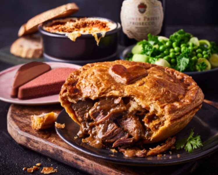 M&S meal example A meat pie on a plate with a cut piece, surrounded by broccoli, mashed potatoes, a dish with melted cheese, and a bottle of sparkling wine.