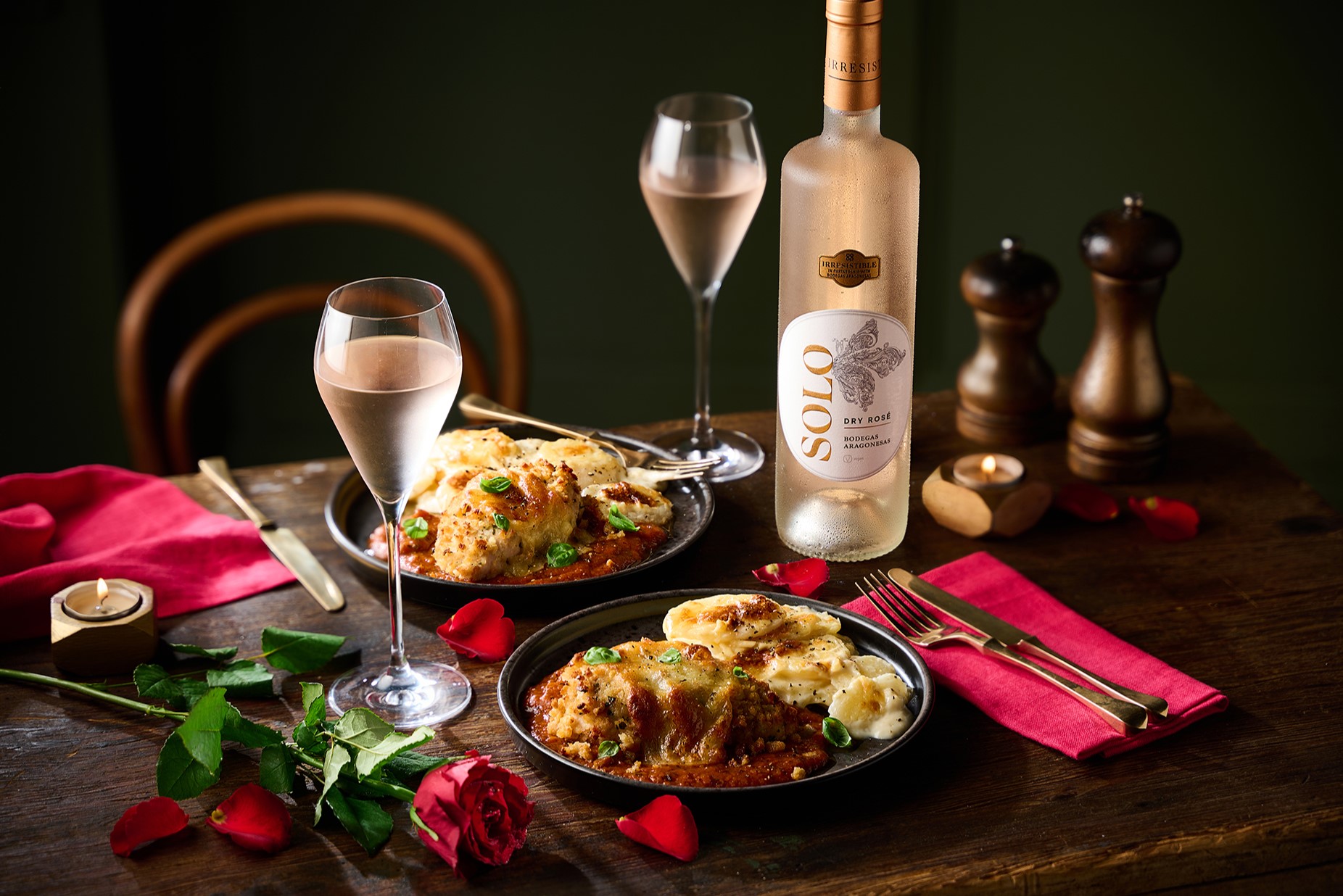 Coop meal deal Two plates of lasagna, a bottle of white wine, wine glasses, red napkins, roses, candles, and salt and pepper shakers on a wooden table.